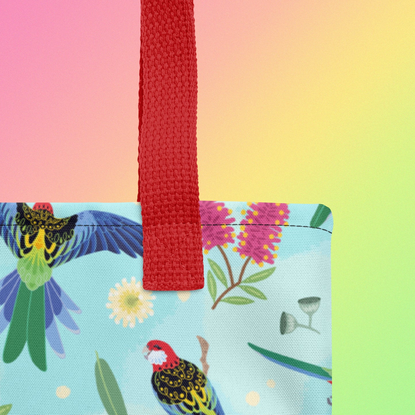 Close up of a tote bag with red handles and an eastern rosella parrot pattern on a gradient pink, yellow and green background