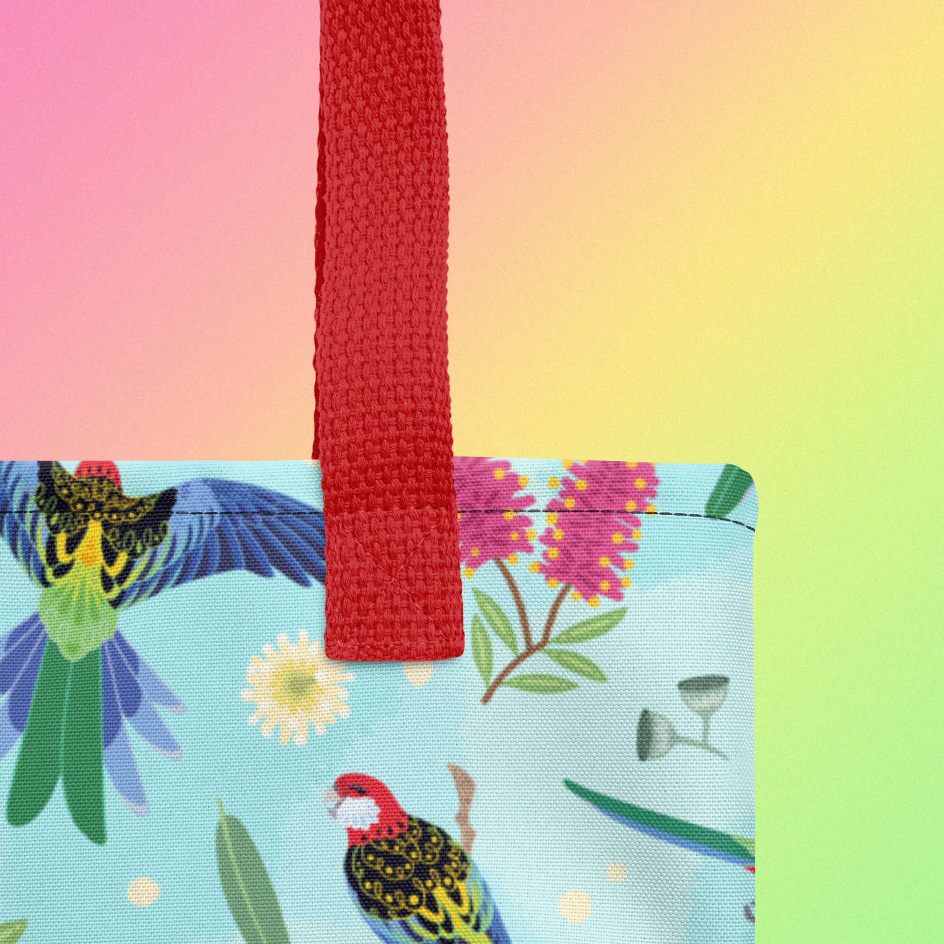 Close up of a tote bag with red handles and an eastern rosella parrot pattern on a gradient pink, yellow and green background