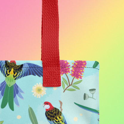 Close up of a tote bag with red handles and an eastern rosella parrot pattern on a gradient pink, yellow and green background