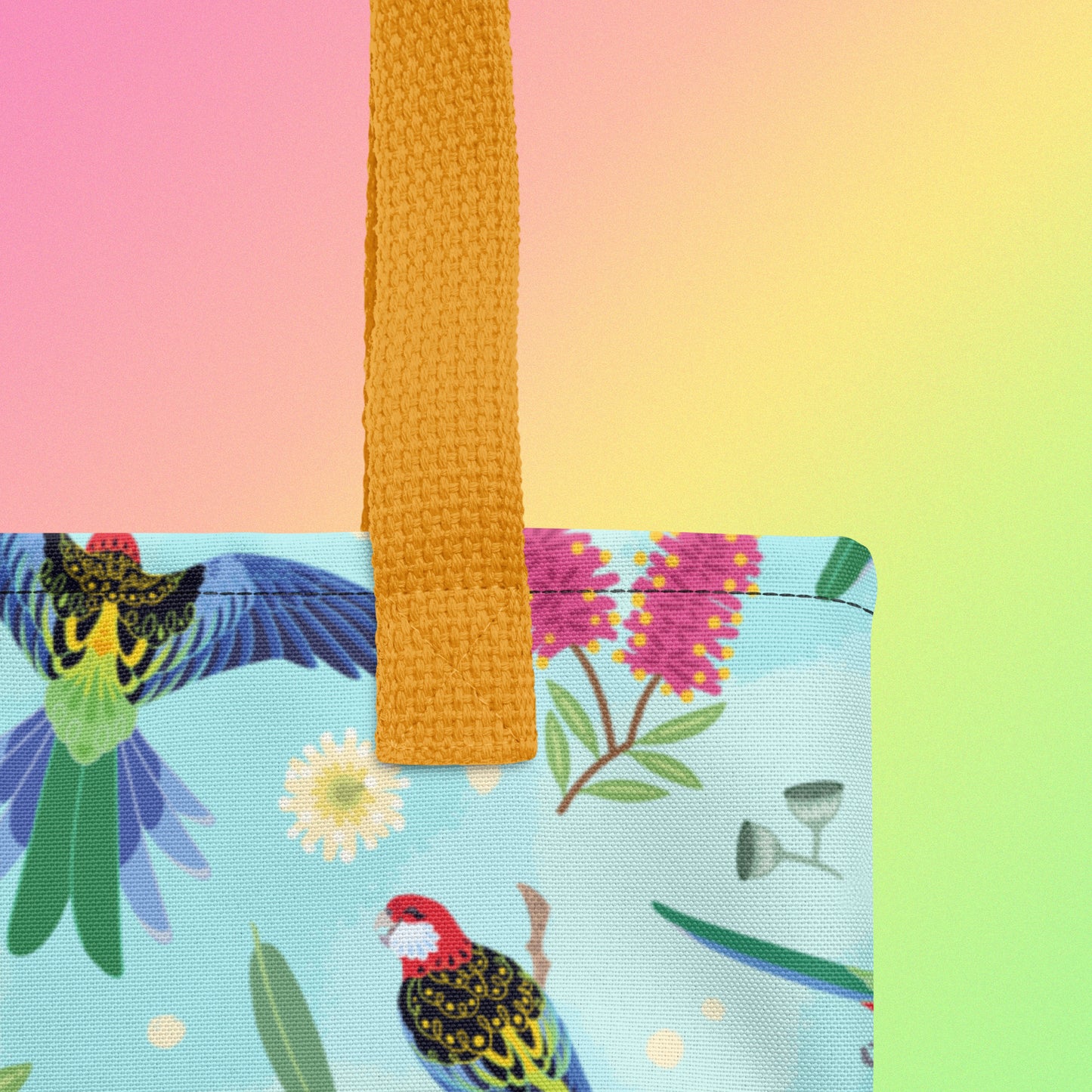 Close up of a tote bag with yellow handles and an eastern rosella parrot pattern on a gradient pink, yellow and green background