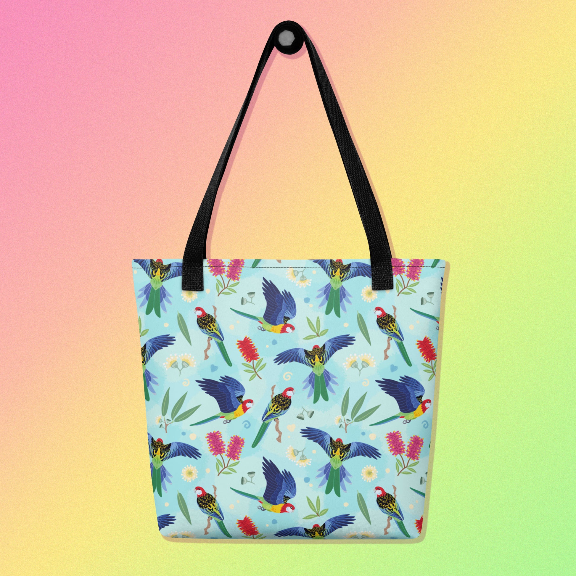 Tote bag with black handles and an eastern rosella parrot pattern on a gradient pink, yellow and green background