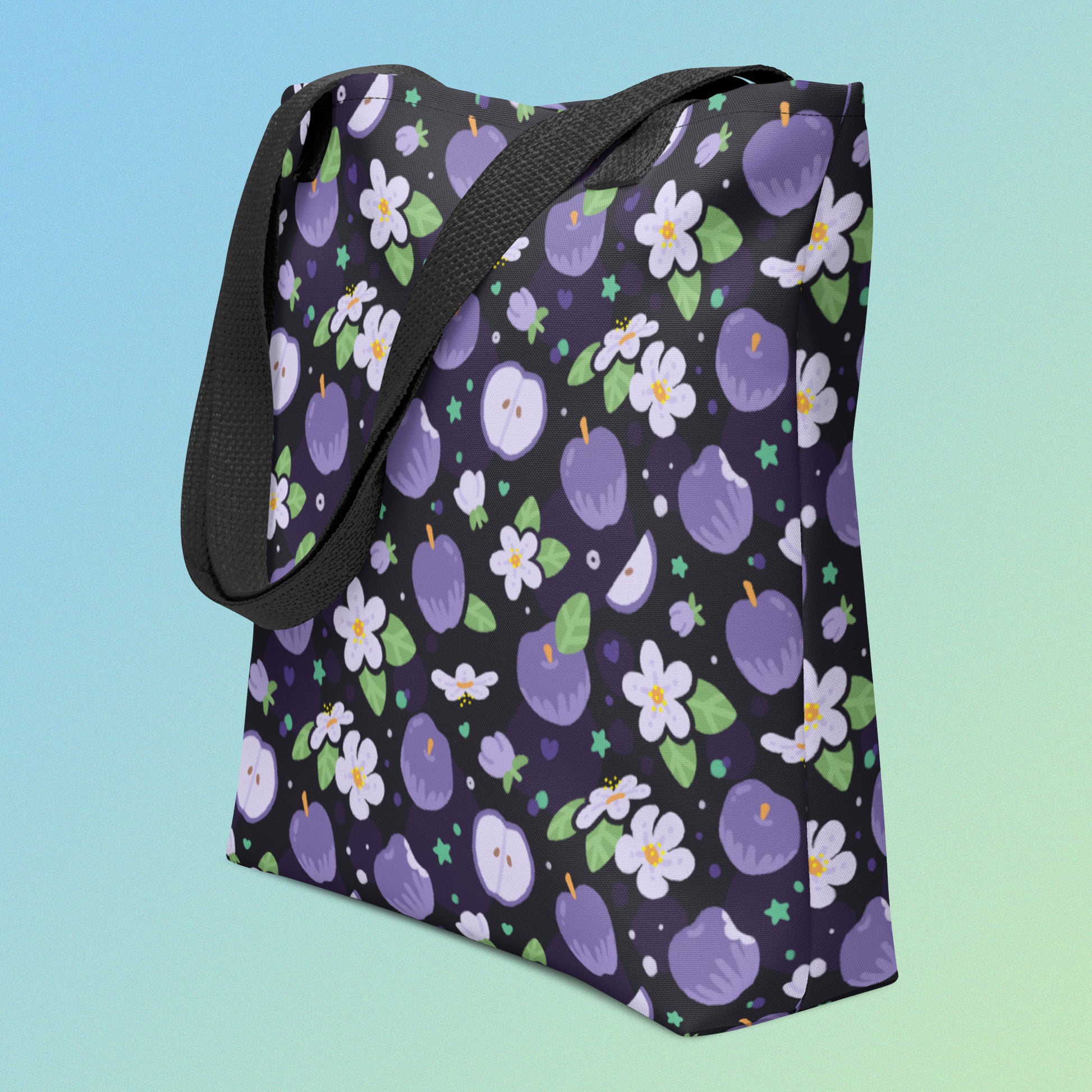 Tote bag with a black strap and a purple apple and flower pattern on an angle on a gradient blue and green background 