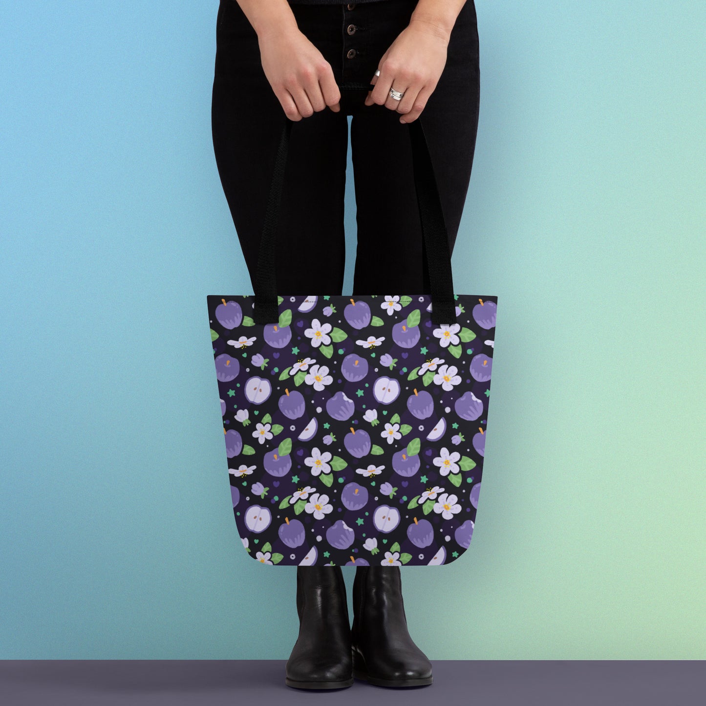 Person holding a tote bag with a black strap and purple apple and flower pattern on a gradient blue and green background