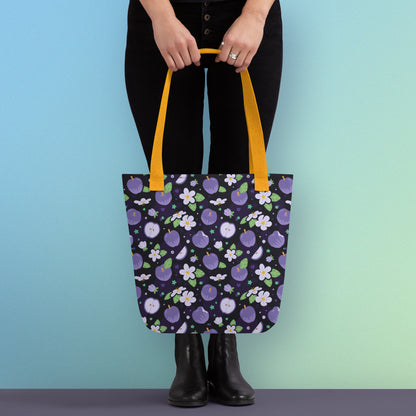 Person holding a tote bag with a yellow strap and purple apple and flower pattern on a gradient blue and green background