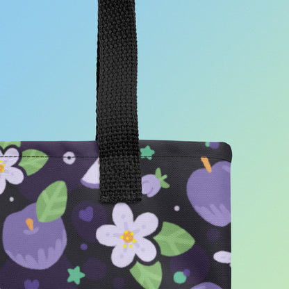 Close up shot of a tote bag with a black strap and purple apple and flower pattern on a gradient blue and green background