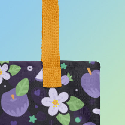 Close up of a tote bag with a yellow strap and purple apple and flower pattern on a gradient blue and green background