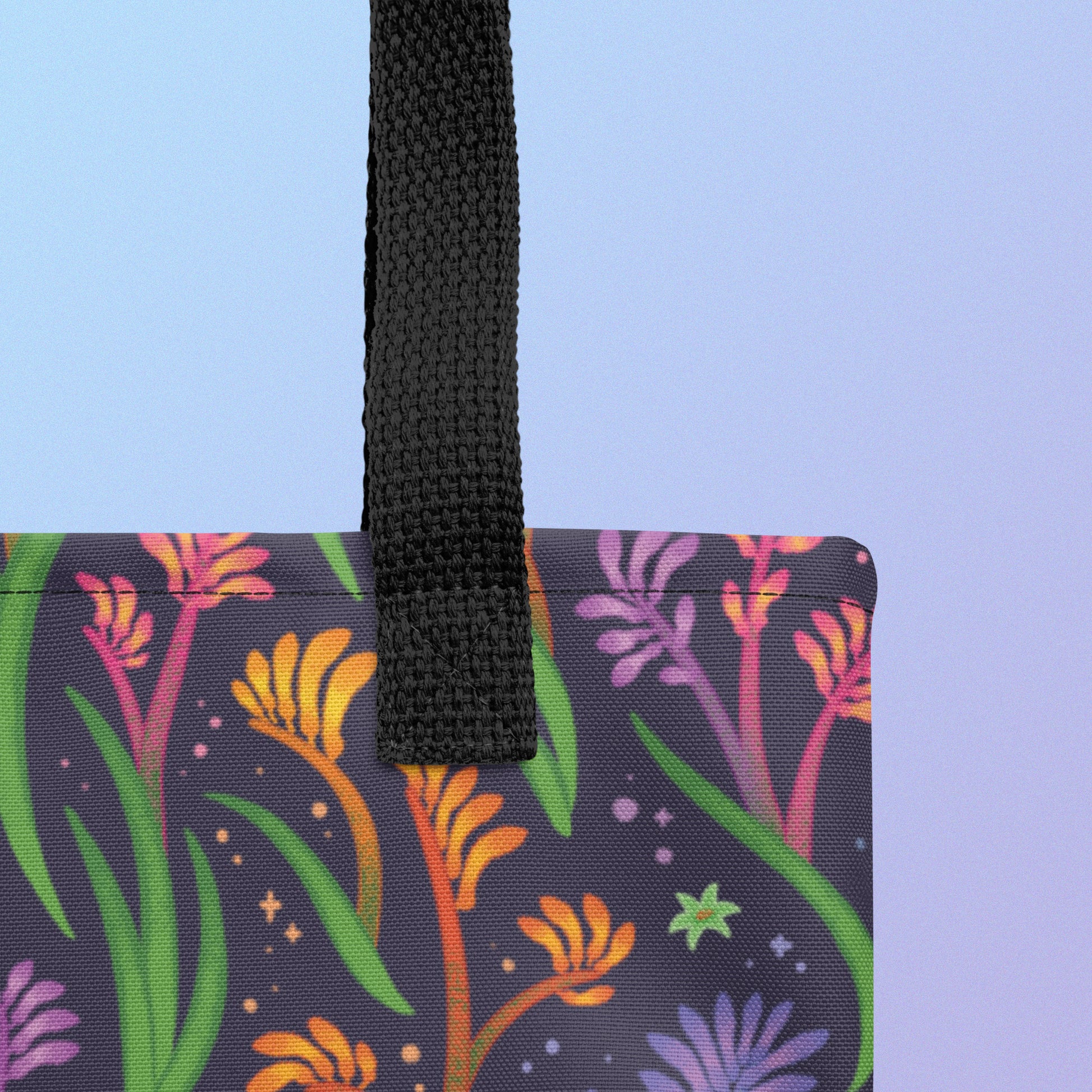Close up of a tote bag with a black strap and a floral rainbow kangaroo paw pattern on a gradient blue and purple background.