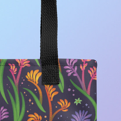 Close up of a tote bag with a black strap and a floral rainbow kangaroo paw pattern on a gradient blue and purple background.
