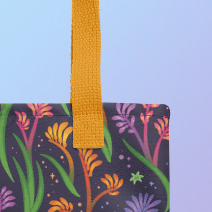 Close up of a tote bag with a yellow strap and a floral rainbow kangaroo paw pattern on a gradient blue and purple background.