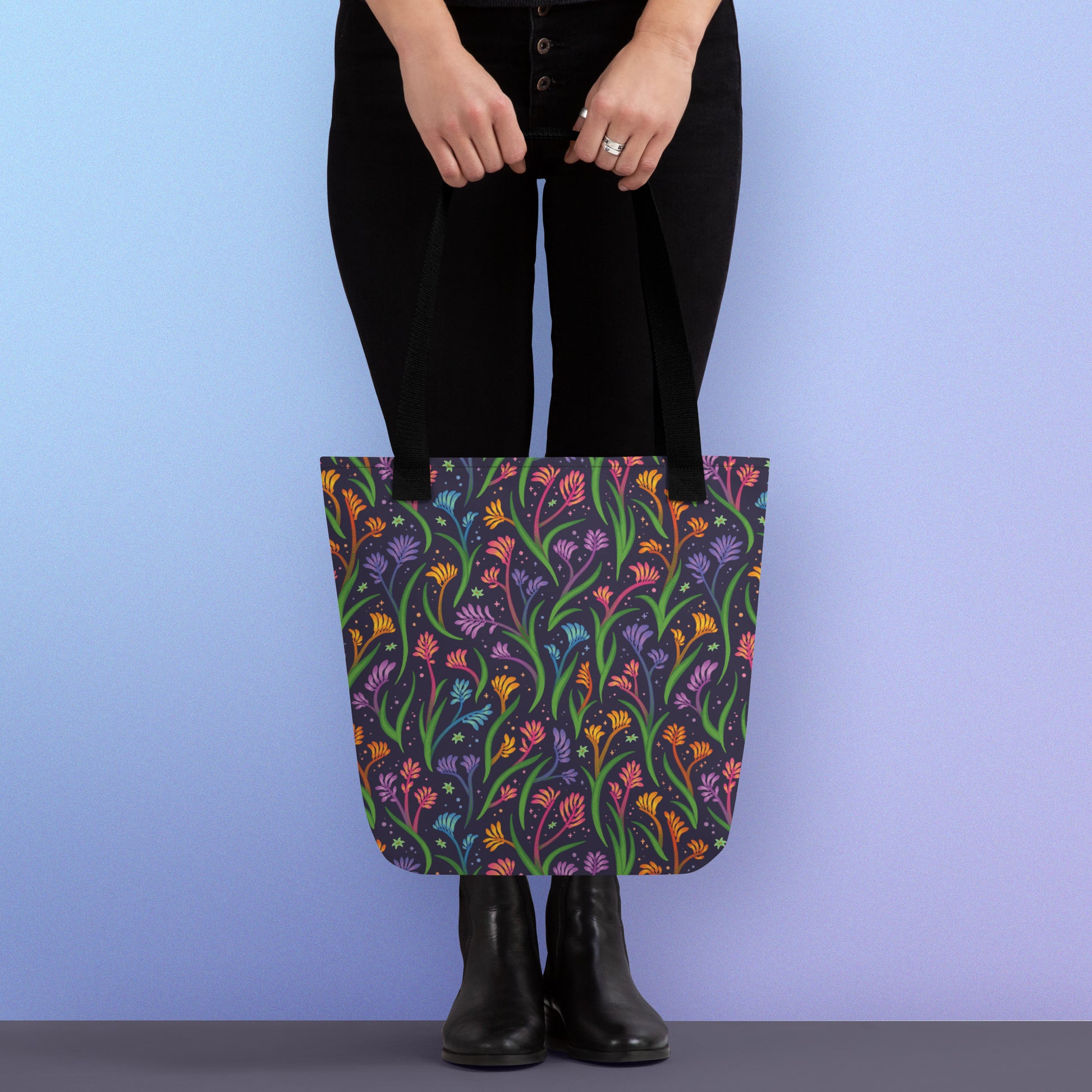 Person holding a tote bag with a black strap and a floral rainbow kangaroo paw pattern on a gradient blue and purple background.