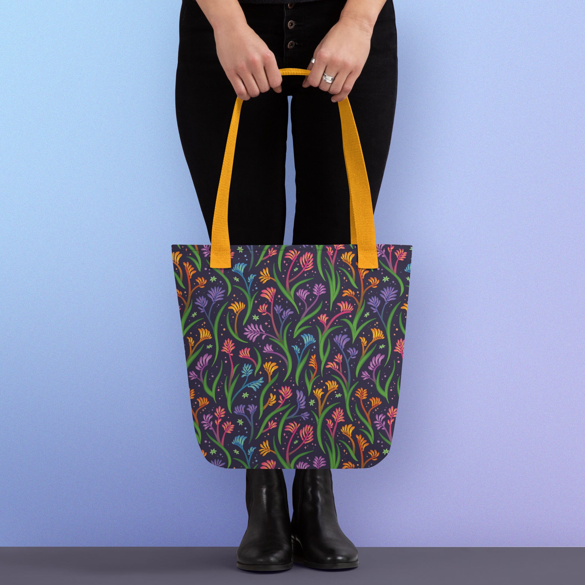 Person holding a tote bag with a yellow strap and a floral rainbow kangaroo paw pattern on a gradient blue and purple background.