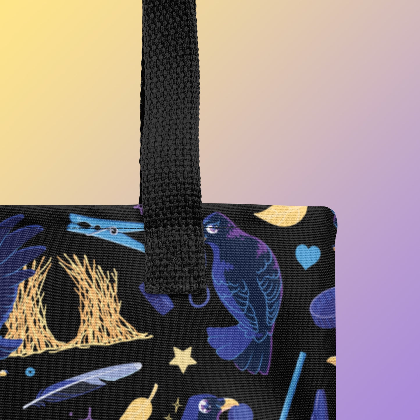 Close up of a tote bag with black handles and a black/blue/yellow satin bowerbird pattern pattern on a yellow and purple gradient background
