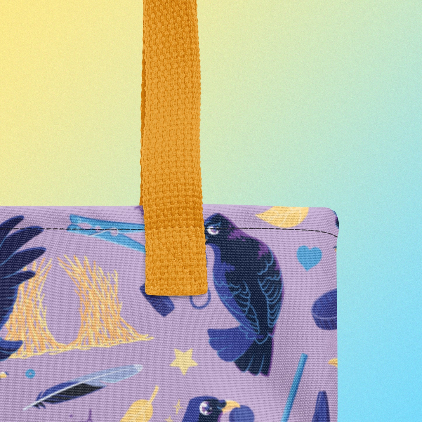 Close up of a tote bag with yellow handles and a purple/blue/yellow satin bowerbird pattern on a yellow and blue gradient background
